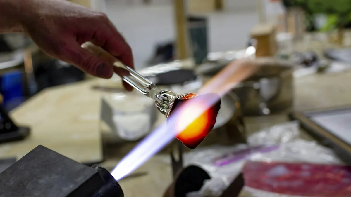Mãos seguram uma vareta de vidro na chama de um maçarico de bancada, com uma gota de vidro incandescente a formar-se na ponta